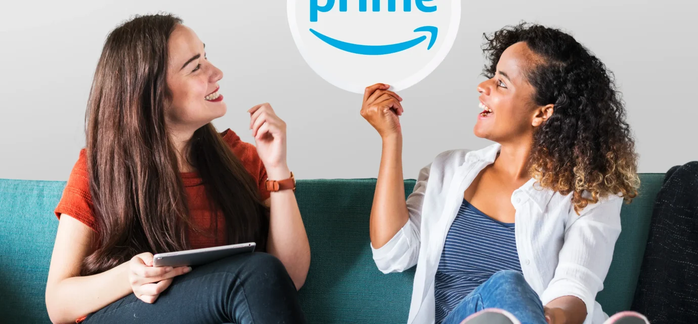 Two young women sitting on a couch, with one holding up a Prime logo sign to discuss amazon prime settlement automatic refunds.