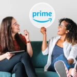 Two young women sitting on a couch, with one holding up a Prime logo sign to discuss amazon prime settlement automatic refunds.