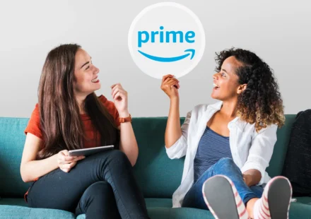 Two young women sitting on a couch, with one holding up a Prime logo sign to discuss amazon prime settlement automatic refunds.