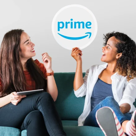 Two young women sitting on a couch, with one holding up a Prime logo sign to discuss amazon prime settlement automatic refunds.