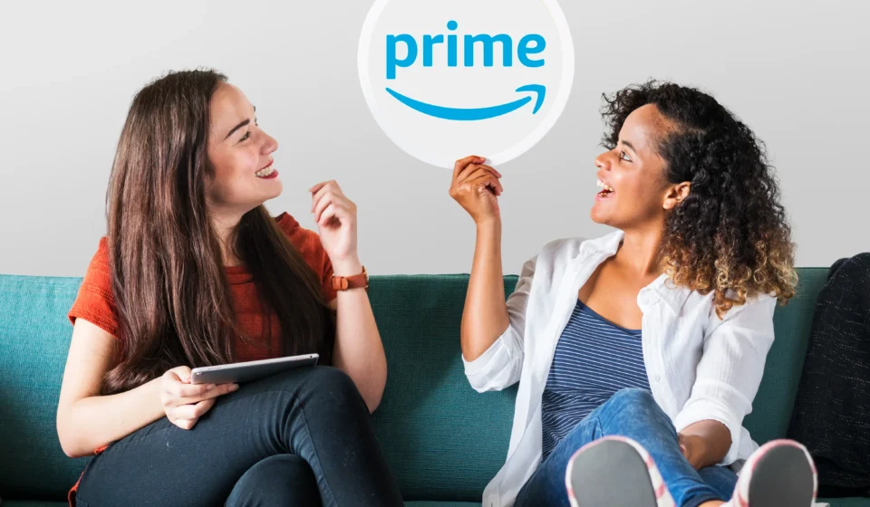 Two young women sitting on a couch, with one holding up a Prime logo sign to discuss amazon prime settlement automatic refunds.