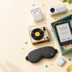 Flat-lay of gadget gift ideas including earbuds, a mini speaker, a portable projector and an e-reader beside a gift box